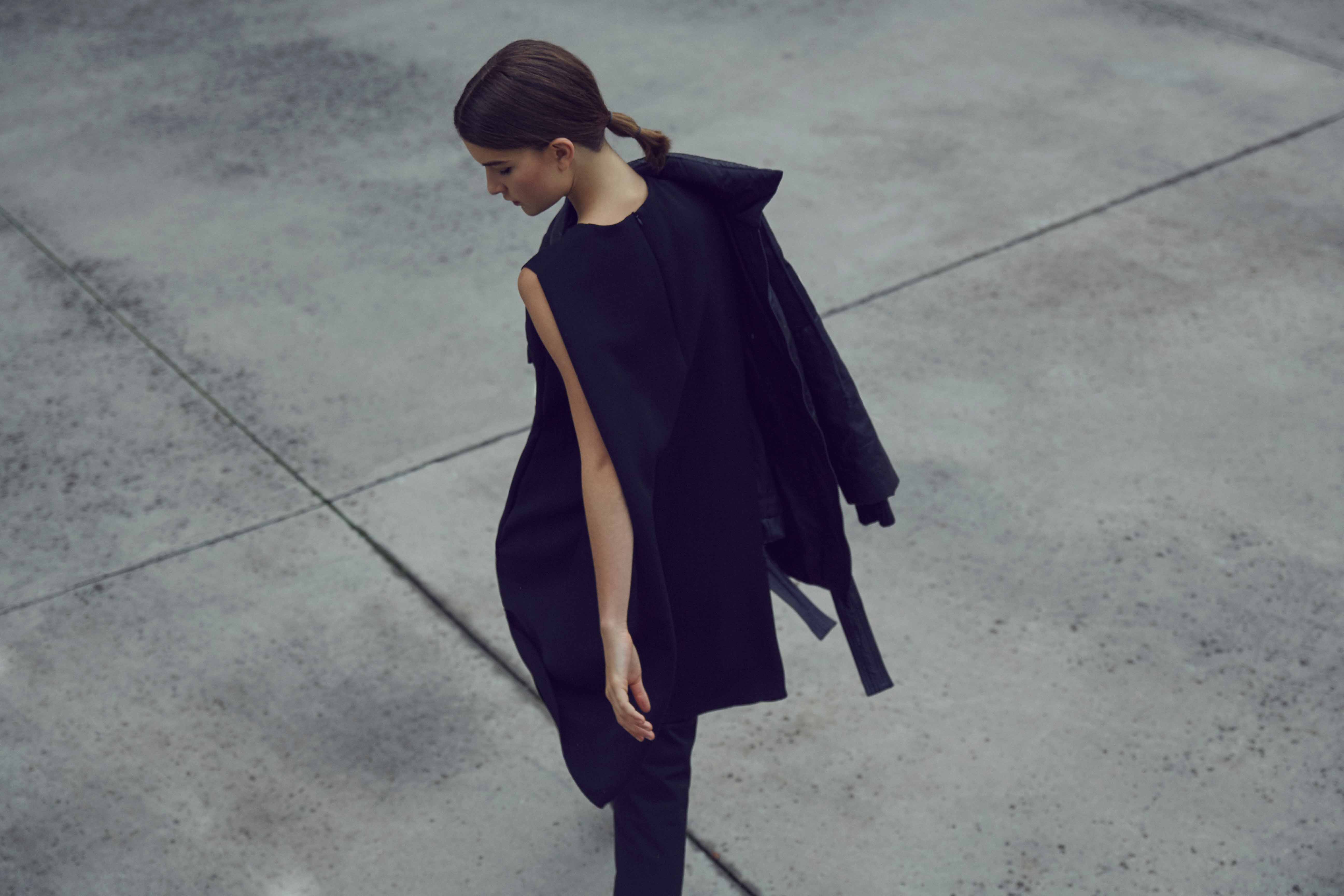 A woman model in a black outfit strides across a concrete pathway. The Shelter Creative - Adolfo Dominguez